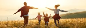 Zwei Eltern mit weit ausgebreiteten Armen und ihre Kinder mit in die Höhe gehaltenen Armen laufen über eine Bergwiese in der Abendsonne.