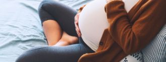 Eine schwangere Frau sitzt auf einem Bett und hält liebevoll eine Hand auf ihren Bauch